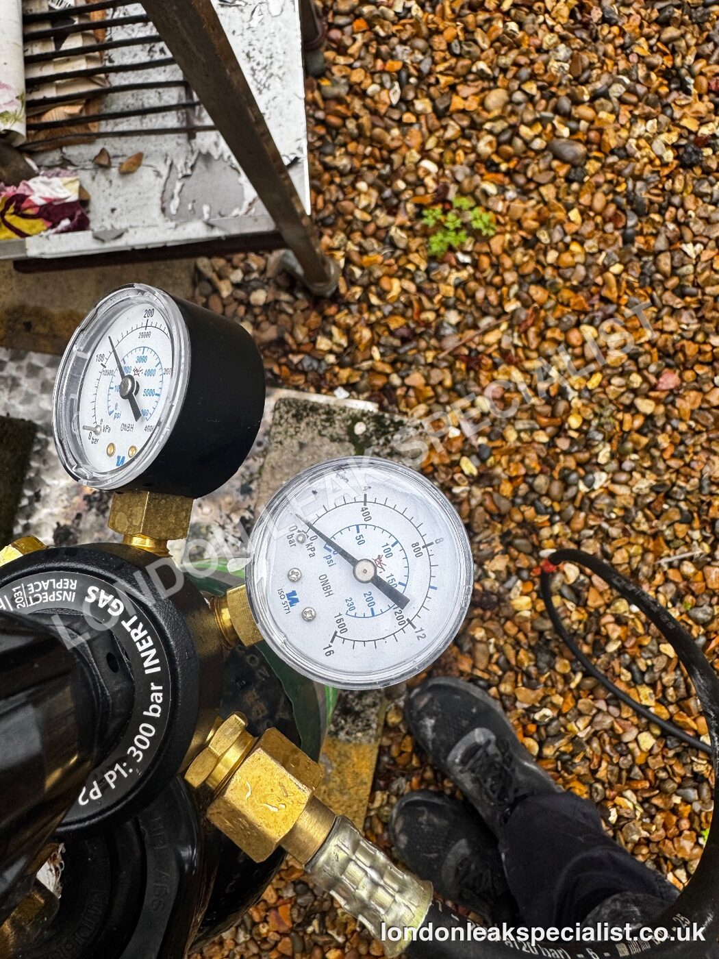 Two pressure gauges on a regulator connected to a hose, resting on gravel, used for a tracer gas test