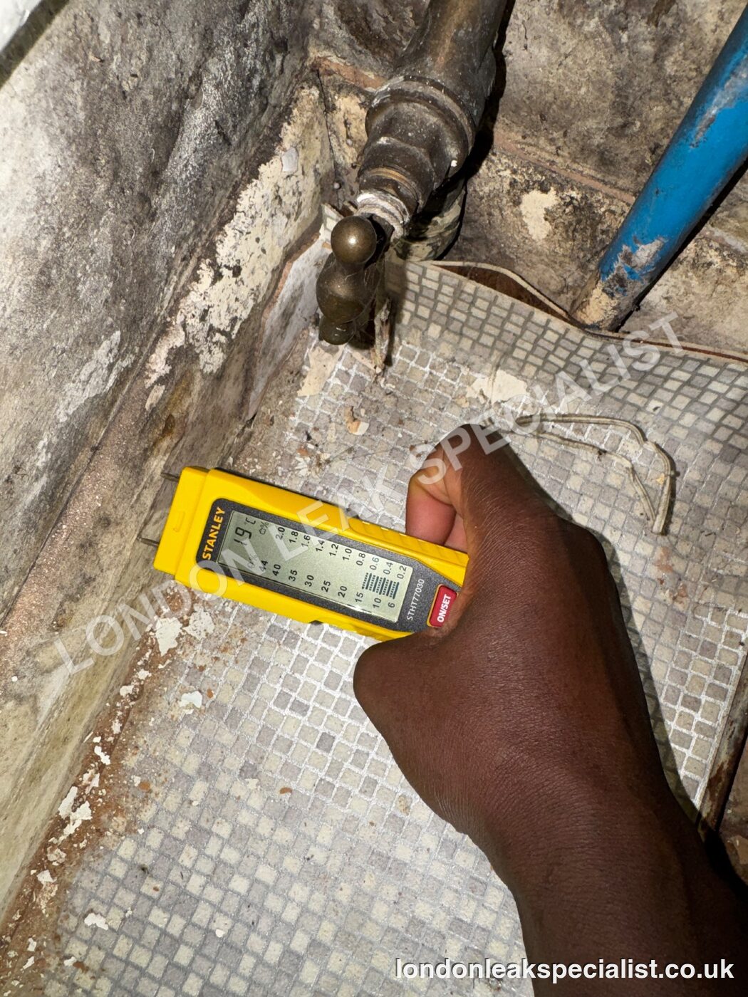 A yellow handheld moisture meter held against a tiled floor edge beside copper and blue plastic pipework
