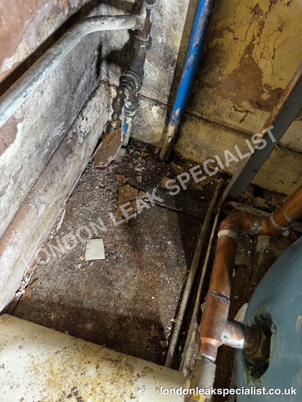 Copper and blue pipework running down a wall into a stained, boxed-in floor area